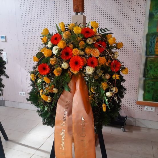 Ein Kranz mit gelben und orangefarbenen Blumen ist an einer weißen Wand ausgestellt. Ein Band mit der Aufschrift 'liebervoller Erinnerung' ist angebracht. Er steht auf einem gefliesten Boden.