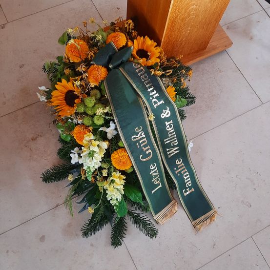 Ein Trauerkranz mit gelben und weißen Blumen, einer grünen Schleife mit goldener Schrift 'Familie Grijse & Wallner', liegt auf einem gefliesten Boden. Dahinter befindet sich eine Holzkanzel.