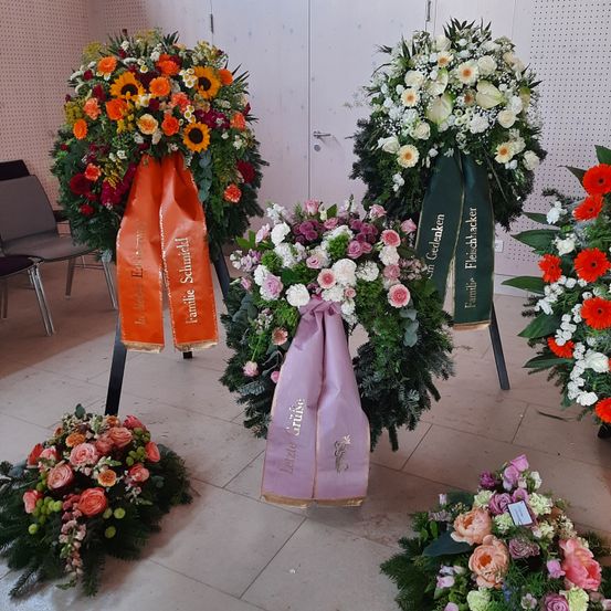 Mehrere Blumenarrangements sind auf einem gefliesten Boden ausgestellt. Es gibt Kränze mit Bändern und einzelne Sträuße in verschiedenen Farben, einschließlich Rosa, Weiß und Orange. Der Hintergrund zeigt eine weiße Wand mit einer Tür.