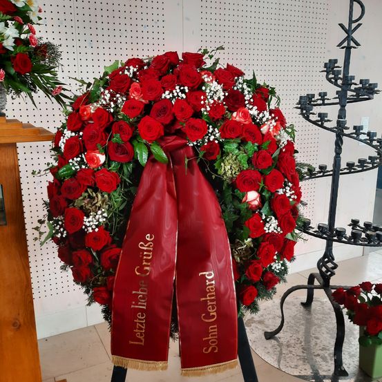 Ein großer Kranz aus roten Rosen mit einem roten Band, auf dem 'Letzte Liebe Gruße Sohn Gerhard' steht, befindet sich auf einem gefliesten Boden. Ein Kerzenständer ist im Hintergrund zu sehen.