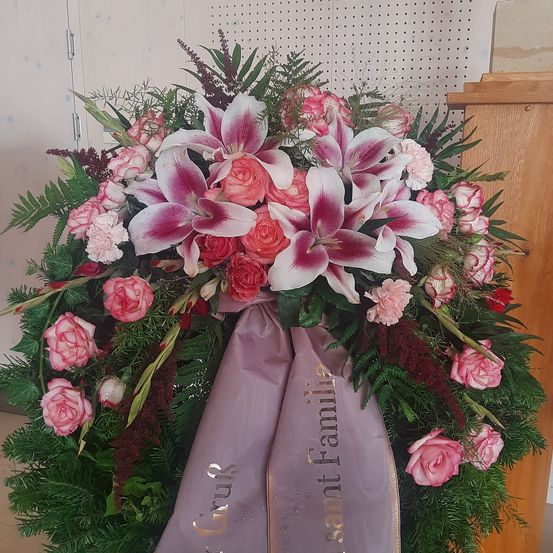 Ein Kranz aus rosa Rosen, Lilien und Grün mit einem lila Band, auf dem 'In liebevoller Erinnerung' steht, wird vor einem weißen Hintergrund gezeigt.