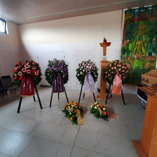 Ein Innenraum mit mehreren Blumenkränzen auf Ständern, einem Holzkreuz und einem Gemälde an der Wand. Stühle und ein hölzerner Podest sind ebenfalls vorhanden.