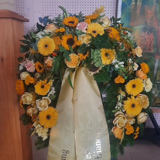 Ein kreisförmiger Blumenkranz aus verschiedenen Blumen wie Rosen, Gänseblümchen und anderen Blüten, mit einem goldenen Band, auf dem 'ea Blanca' steht.