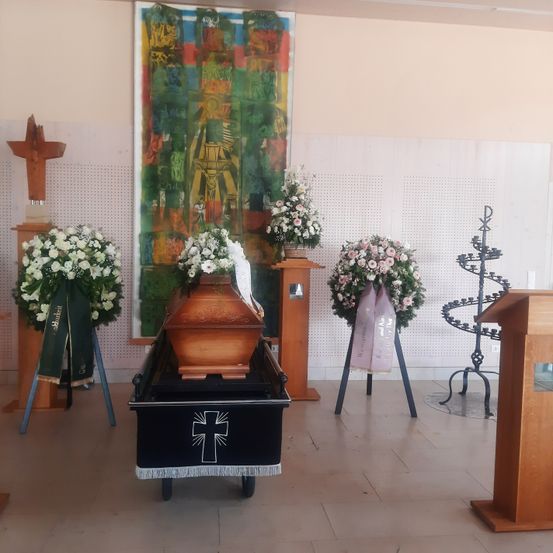 Eine Beerdigungsszene mit einem hölzernen Sarg, weißen und rosa Blumenarrangements und religiösen Symbolen. Ein farbenfrohes Gemälde und ein Kerzenhalter sind im Hintergrund.