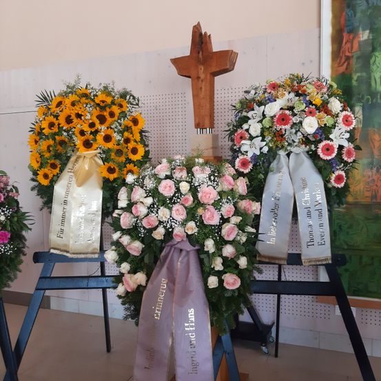 Drei Blumenkränze mit Bändern sind auf Ständern ausgestellt. Einer hat Sonnenblumen, ein anderer hat rosa und weiße Rosen und der dritte ist eine Mischung aus weißen und rosa Blumen. Ein hölzernes Kreuz steht hinter dem mittleren Kranz.