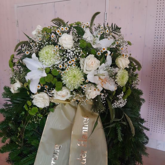 Ein großes Blumenarrangement mit weißen Rosen, grünen Blättern und Gänseblümchen steht vor einem rosa Hintergrund mit einem goldenen Band, das 'In lieber Erinnerung' besagt.