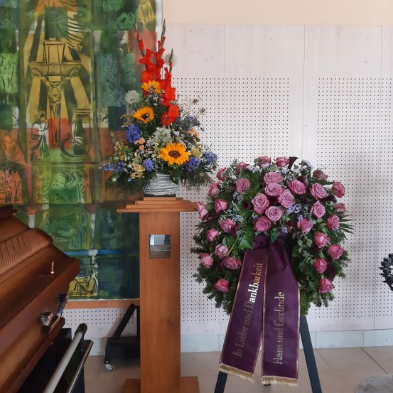 Ein Holzständer hält eine farbenfrohe Blumenarrangement neben einem Sarg. Ein Kranz mit rosa Rosen und einem lila Band befindet sich in der Nähe. Der Hintergrund zeigt ein lebhaftes Gemälde mit religiösen Darstellungen.