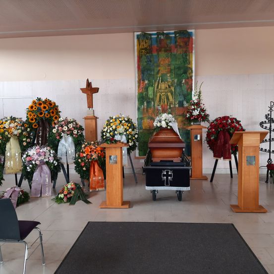 Ein Bestattungsraum mit einem Sarg in der Mitte, umgeben von verschiedenen Blumenarrangements und Holzständern. Der Raum ist mit einer farbenfrohen Wandmalerei und einem Kreuz an der Wand geschmückt.