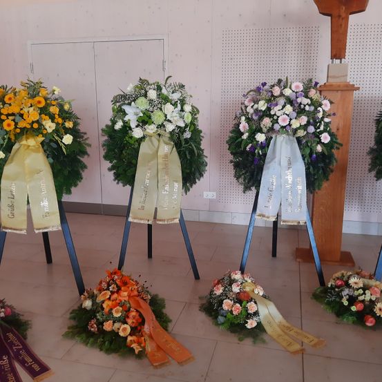 Mehrere Blumenkränze sind auf Ständern in einem Raum ausgestellt. Jeder Kranz hat ein Band mit unterschiedlichem Text. Der Raum hat weiße Wände mit schwarzen Punkten und einem hölzernen Podium. Der Boden ist gefliest.