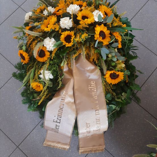 Ein Kranz mit Sonnenblumen, Nelken und Grün, einschließlich einer Schleife mit der Aufschrift 'In liebevoller Erinnerung' und einem Namen darauf.