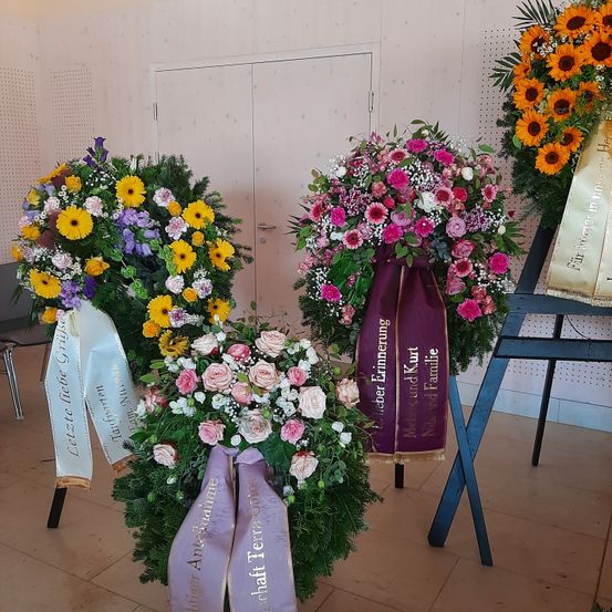 Drei Blumenarrangements sind auf einem gefliesten Boden ausgestellt. Jedes Arrangement hat ein anderes Farbschema, wobei eines gelbe und lila Blumen, ein anderes rosa und weiße Blumen und das dritte eine Mischung aus Farben aufweist. Jedes hat ein Band mit Text.