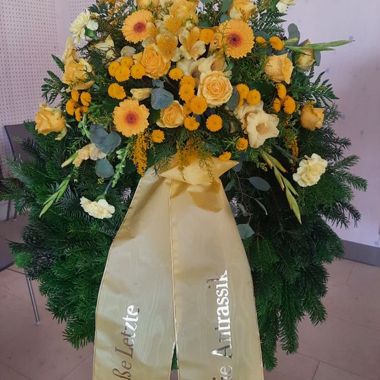 Eine große Blumenarrangement mit gelben Blumen und grünen Blättern. Der Strauß ist kreisförmig mit einem gelben Band, das 'In Erinnerung' steht. Es ist auf einem gefliesten Boden ausgestellt.