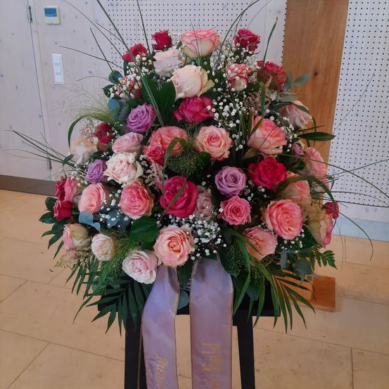 Eine große Blumenarrangement mit Rosen in verschiedenen Farben, darunter rosa, rot und lila, und Babys Atmung. Die Blumen sind auf einem schwarzen Ständer mit zwei lila Bändern angeordnet, und der Hintergrund ist eine Wand mit einem weißen und grauen Muster.