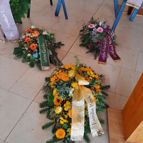 Drei Blumenkränze liegen auf dem Boden. Sie haben grüne und weiße Bänder mit Namen. Es gibt lila Blumen in den Kränzen. Sie liegen auf einem gefliesten Boden.