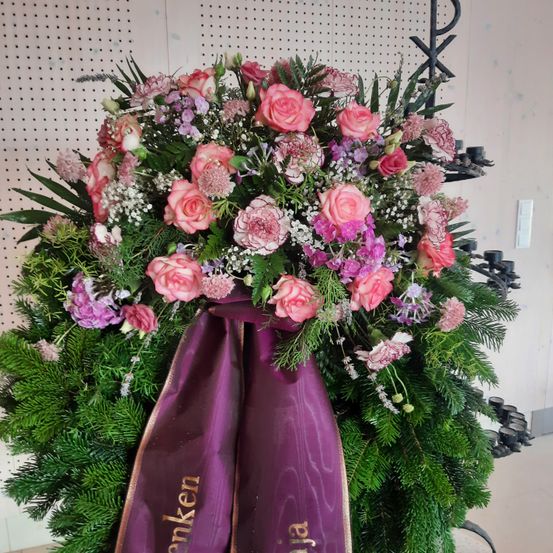 Eine große Blumenarrangement mit rosa und lila Blumen und einem lila Band mit goldenen Buchstaben, die 'Banken' lesen. Das Arrangement ist an einer weißen Wand mit einem Kerzenhalter ausgestellt.