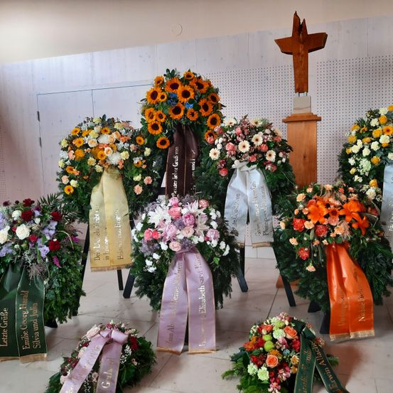 Eine Sammlung von Blumenkränzen, die in einem Raum angeordnet sind, einige mit Bändern und einem Kreuz im Hintergrund. Die Kränze zeigen eine Vielzahl von Blumen, darunter Sonnenblumen, Rosen und Gänseblümchen.