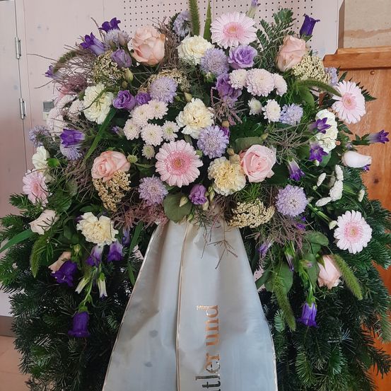 Eine Blumenarrangement mit einer Mischung aus rosa, lila und weißen Blumen. Der Strauß ist mit grünen Blättern und einem weißen Band mit goldener Schrift verziert. Die Blumen sind mit einem Band zusammengebunden.