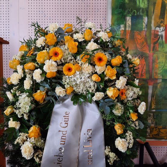 Ein Kranz aus weißen und gelben Blumen, einschließlich Rosen, Nelken und Gänseblümchen, ist an einer Wand mit einem Gemälde ausgestellt. Ein grauer Band mit goldenem Text ist am Kranz befestigt.
