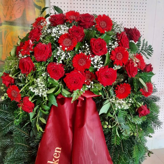 Ein großer Kranz mit leuchtend roten Rosen, Babysatem und grünen Blättern. Eine rote Schleife mit goldener Beschriftung 'Kalken'. Dahinter ist ein weißer Hintergrund mit einem Gemälde.