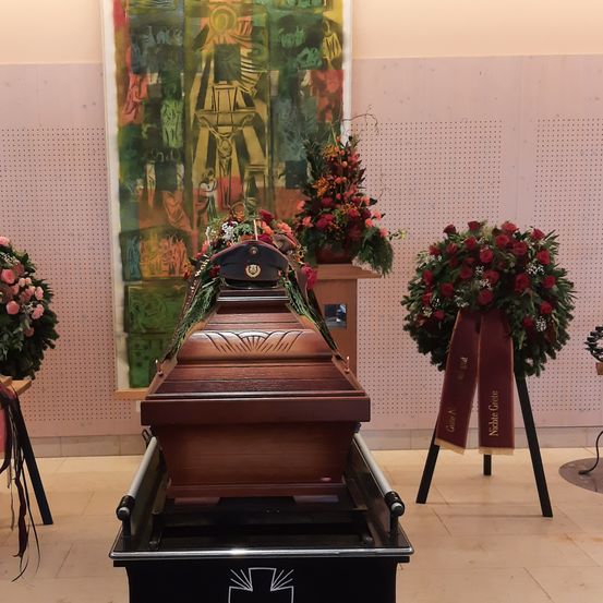 Ein Sarg mit einem Polizeihut auf einem Ständer mit Blumenarrangements. Dahinter befindet sich ein Gemälde.