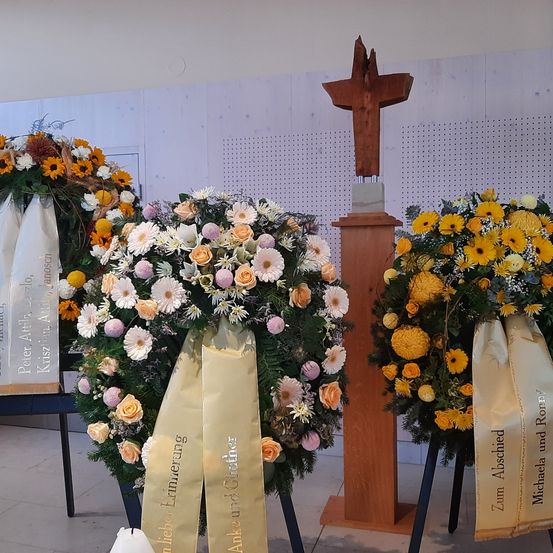 Drei Blumenkränze sind in einem Raum auf Ständern ausgestellt, jeder mit Bändern. Ein Kranz ist gelb und weiß, ein anderer gelb und orange, und der dritte weiß und gelb. Hinter den Kränzen steht ein Holzkreuz.