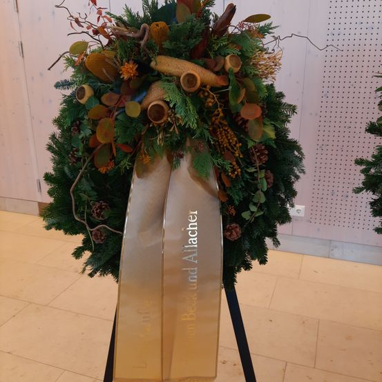 Ein Kranz aus verschiedenen Naturmaterialien wie grünen Blättern, Tannenzapfen und getrockneten Früchten steht auf einem dekorativen Ständer. Der Kranz hat ein goldenes Band mit dem Text 'Laurier et Allaché'.
