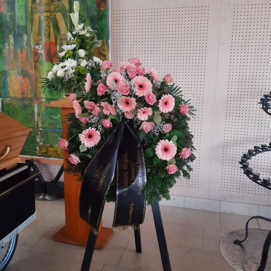 Ein rosa und weißes Blumenarrangement auf einem Ständer mit einem schwarzen Band, das 'in lieber Erinnerung' steht, befindet sich in einem Raum mit weißer Wand und einem Sarg.