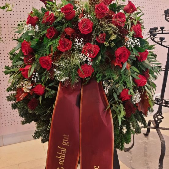 Ein Kranz mit roten Rosen, grünen Blättern und einer roten Schleife mit der Aufschrift 'gut schlaf' steht vor einer rosa Wand.