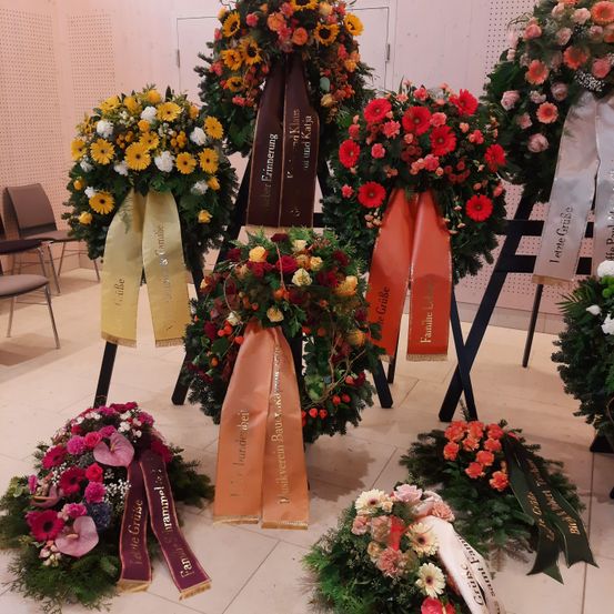 Mehrere Kränze mit Bändern und Blumen, darunter Rosen, Gänseblümchen und andere Blumen, sind auf einem Holzständer ausgestellt. Der Hintergrund besteht aus einer Wand mit einer weißen Tür und einigen Stühlen.