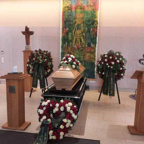 Ein hölzerner Sarg steht auf einer schwarzen Plattform, umgeben von Kränzen und Blumengestecken. Ein hölzerne Kreuz auf einem Podest zur Linken. Ein farbenfrohes Wandbild an der Wand hinter dem Sarg.