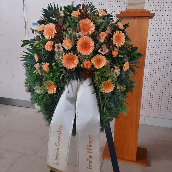 Ein Kranz mit orangefarbenen Blumen und grünen Blättern, mit einem weißen Band gebunden, steht auf einem Holzpodium. Das Band trägt die Aufschrift 'In lieber Gedenken' und 'Familie Pillinger'. Der Hintergrund zeigt eine weiße Wand und einen gefliesten Boden.