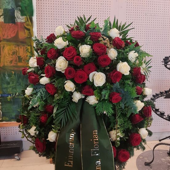 Ein Kranz aus roten und weißen Rosen mit grünen Blättern und einem grünen Band, das 'Einenen Florian' steht, ist in einem Raum ausgestellt.