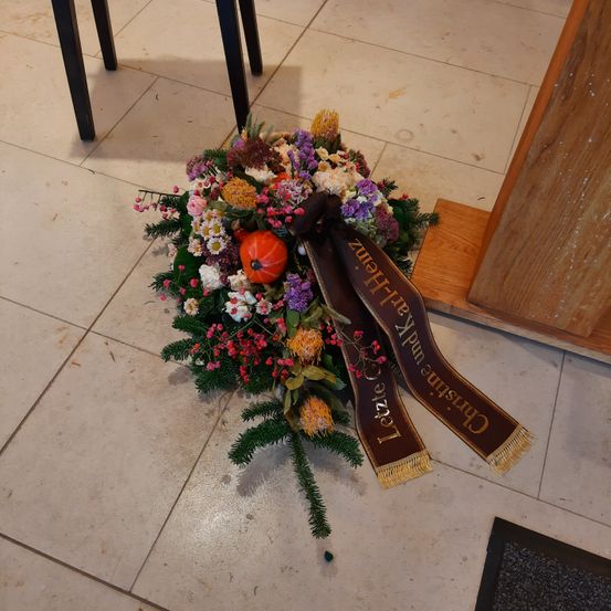 Ein Blumenarrangement mit verschiedenen Blumen, darunter Rosen, Gänseblümchen und Beeren, liegt auf einem gefliesten Boden. Eine Schleife mit der Aufschrift 'Letzte Ehre Dankbarkeit'. Ein kleiner Kürbis ist ebenfalls enthalten.