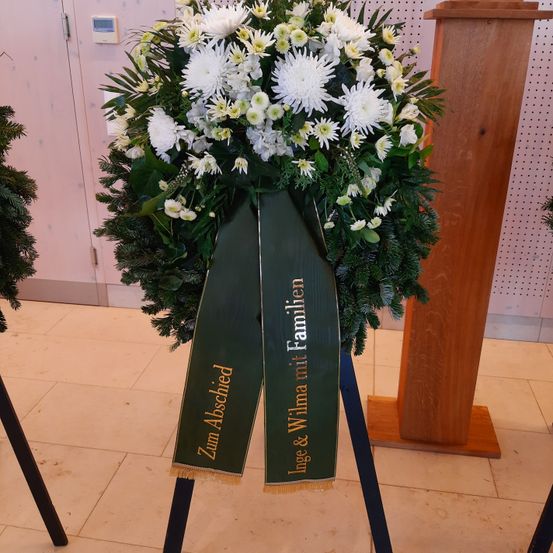 Ein Kranz aus weißen Blumen mit einem grünen Band, das Zum Abschied steht. Er steht auf einem Ständer vor einer rosa Wand.