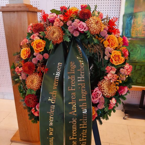 Ein Kranz mit einem grünen Band, auf dem 'Freunde Andrea Fredrich Heinig' steht, wird auf einem Holzständer vor einer weißen Wand ausgestellt. Der Kranz ist farbenfroh mit Rosen, Chrysanthemen und anderen Blumen.