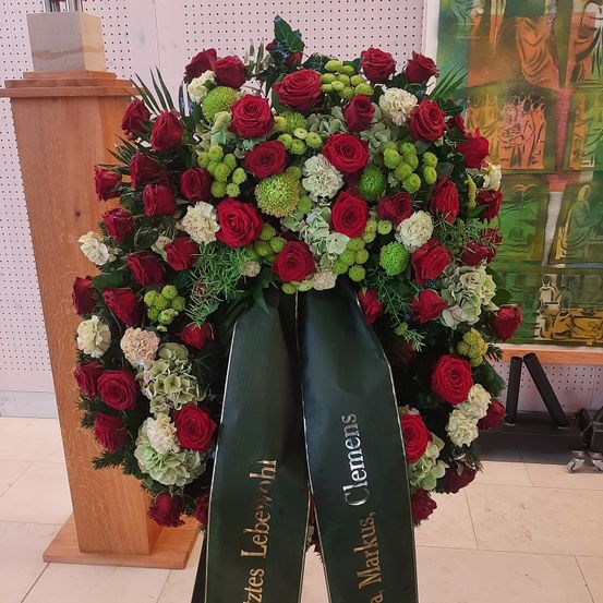 Ein großer Blumenkranz aus roten, weißen und grünen Blumen mit einem grünen Band, auf dem Clemens und Markus steht. Er steht auf einem gefliesten Boden.