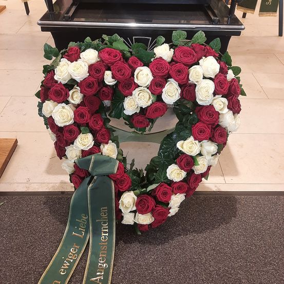 Ein herzförmiger Kranz aus roten und weißen Rosen mit grünen Blättern und einer grünen Schleife mit den Worten 'ewiger Liebe' liegt auf einem Marmorboden.