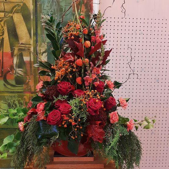 Eine farbenfrohe Blumenkomposition mit roten Rosen, orangefarbenen Beeren und grünen Blättern wird in einem roten Topf vor einem weißen Hintergrund mit einem gemusterten Wand gezeigt.