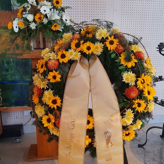 Ein dekorativer Kranz aus Sonnenblumen und grünen Blättern mit einem goldenen Band, das 'In lieber Grus' sagt, wird auf einem Holzständer vor einer weißen Wand ausgestellt.