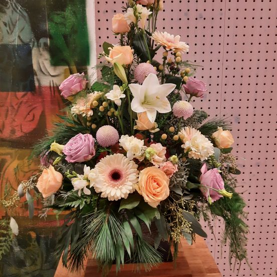 Eine große Blumenarrangement mit einer Mischung aus rosa, weißen und gelben Blumen und grünen Blättern wird auf einem Holztisch vor einer rosa Wand mit einem Gemälde ausgestellt.