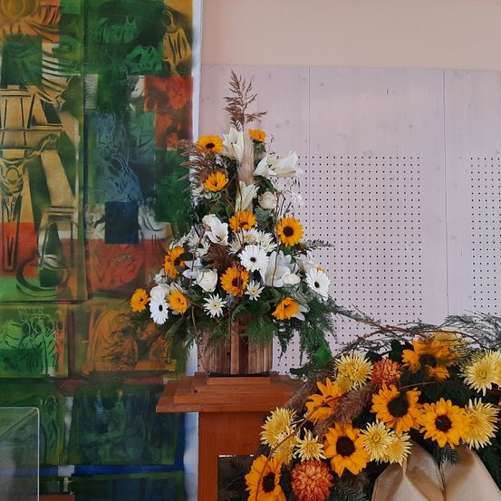 Eine Blumenarrangement aus weißen und gelben Blumen steht auf einem Holztisch. Dahinter befindet sich ein farbenfrohes abstraktes Gemälde.