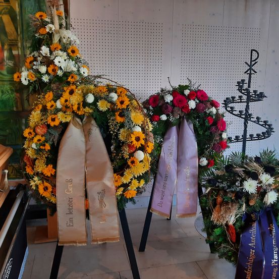 Drei Kränze, zwei mit den Bändern 'Ein letzter Gruß' und 'Du wirst uns unsichtbar', sind vor einer weißen Wand ausgestellt. Die Kränze bestehen aus verschiedenen Blumen, darunter Sonnenblumen, Gänseblümchen und rote Rosen.