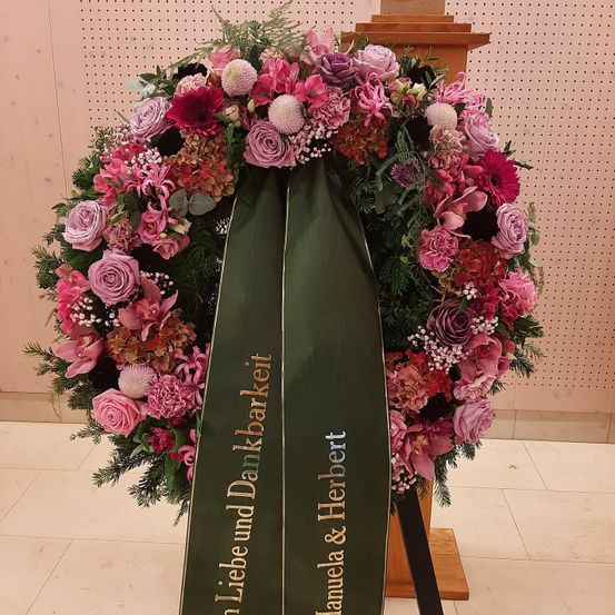 Ein Kranz mit rosa Blumen und einem grünen Band, mit dem Namen Liebe und Dunkelbarkei, wird auf einem Holzständer in einem Raum ausgestellt.