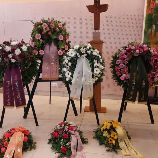 Mehrere Blumenkränze sind auf Ständern in einem Raum ausgestellt. Die Kränze haben Bänder und Blumen. Ein Kreuz steht auf einem Ständer hinter den Kränzen.