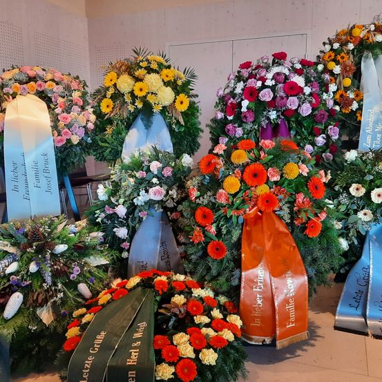 Eine Sammlung von Kränzen und Blumengestecken in verschiedenen Farben wird ausgestellt, einige mit blauen Bändern mit Text. Die Blumen umfassen Rosen, Nelken und Gänseblümchen.