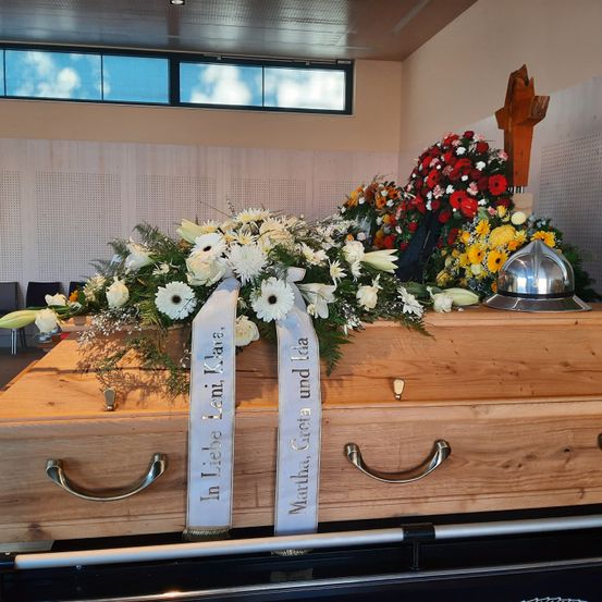 Ein hölzerner Sarg, geschmückt mit Blumen und einem silbernen Helm, ist in einem Raum mit Fenstern ausgestellt. Hinter dem Sarg steht ein hölzernes Kreuz und weitere Blumen.