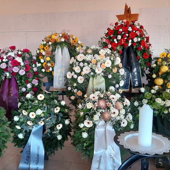 Eine Sammlung von Blumenkränzen mit Bändern in verschiedenen Farben, die an einer Wand ausgestellt sind. Jeder Kranz hat eine andere Blumenanordnung, und einige haben ein Kreuz oben. Eine Kerze ist auf einer Spitze auf einem kleinen Tisch platziert.