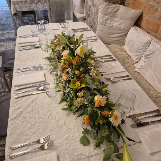 Ein Tisch für eine Mahlzeit mit Besteck, Gläsern und einem Blumenarrangement. Im Hintergrund befinden sich Kissen und eine Ziegelwand.