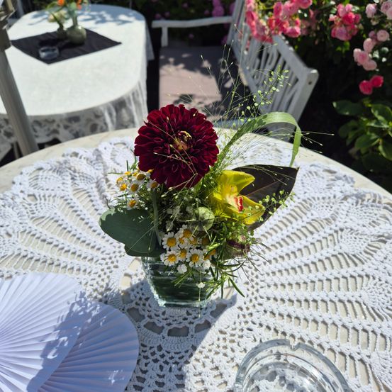 Ein Tisch mit einem weißen Spitzendeckchen hat eine Vase mit einer bunten Blumenarrangement. In der Nähe befinden sich weitere Tische mit weißen Tischdecken und eine weiße Bank.