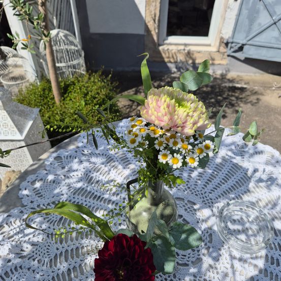 Ein Blumenarrangement in einer Vase auf einem Tisch mit einer weißen Spitzentischdecke. Im Mittelpunkt steht eine große rosa Blume mit gelben Blütenblättern, umgeben von Gänseblümchen und Grün.